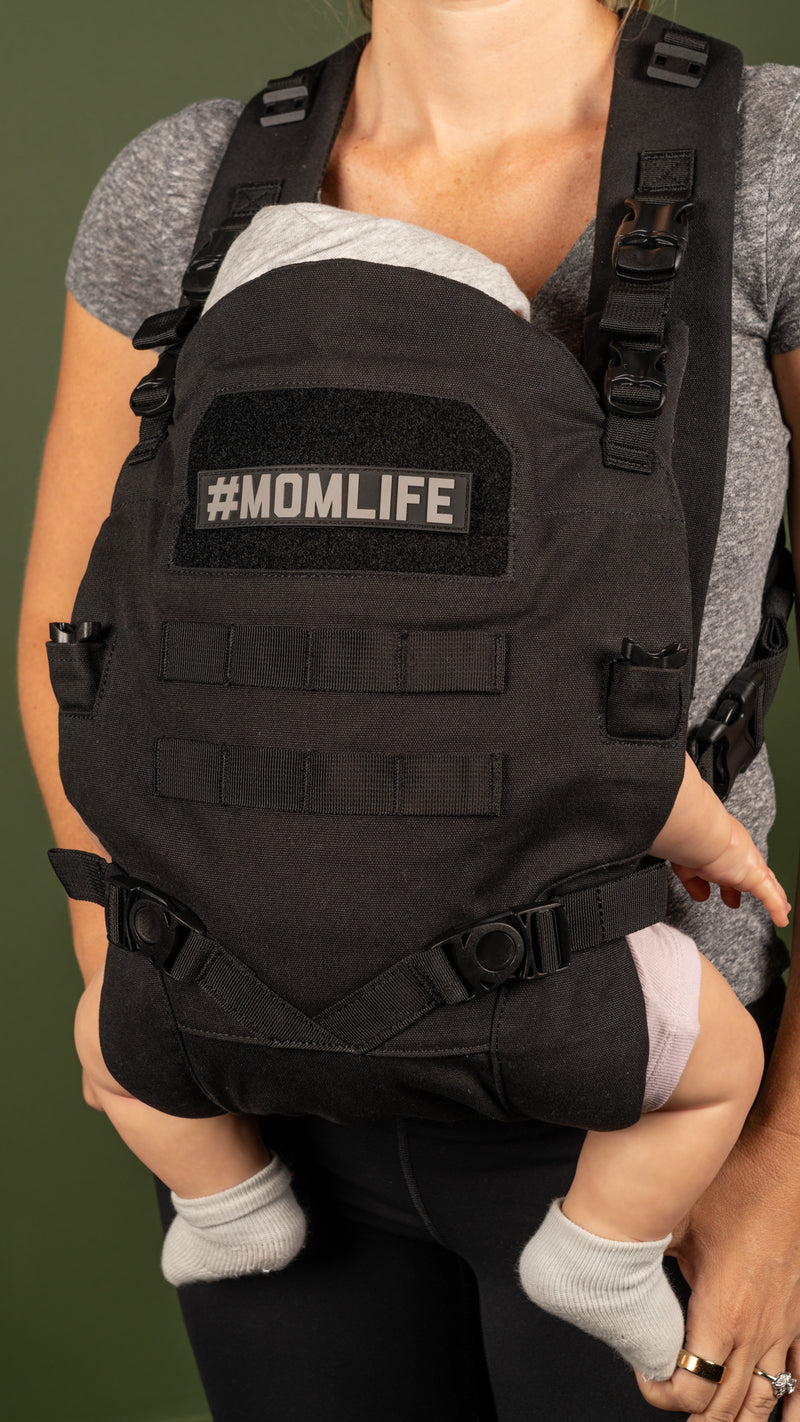 Woman wearing a black, military-style baby carrier with "#MOMLIFE" patch.