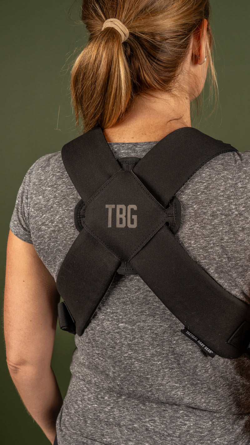 Woman wearing a black, tactical baby carrier with "TBG" logo on a grey background.