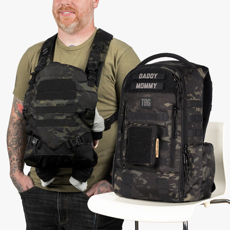 A man wearing a camouflage daypack diaper bag with baby carrier straps, standing next to a matching camouflage diaper bag backpack labeled Daddy and Mommy.