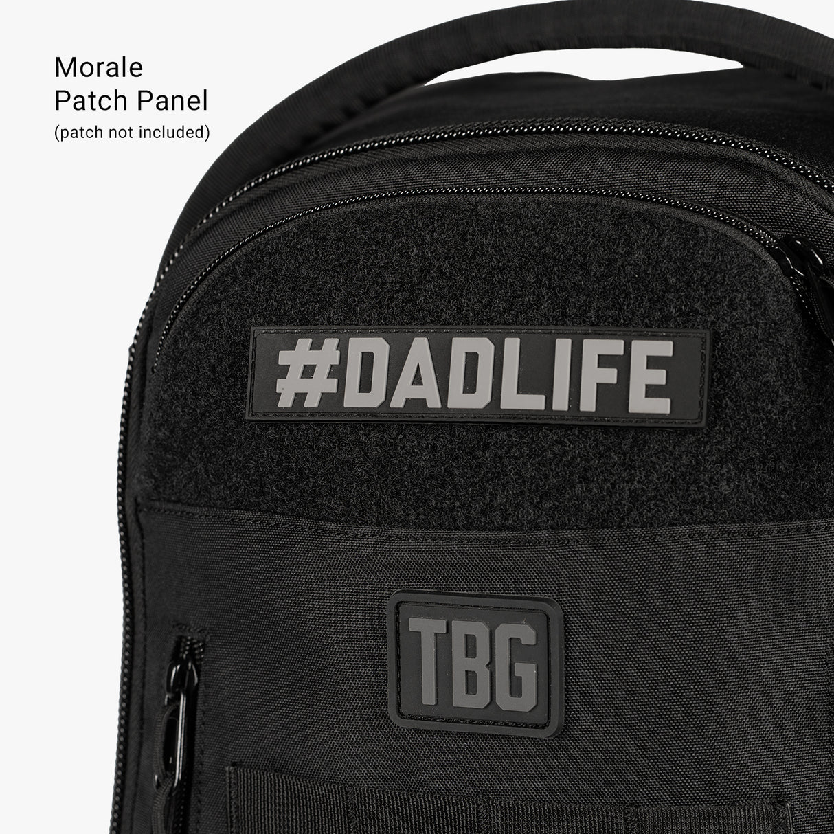 Black daypack diaper bag with patches reading "#DADLIFE" and "TBG," designed for carrying baby essentials and a baby carrier.