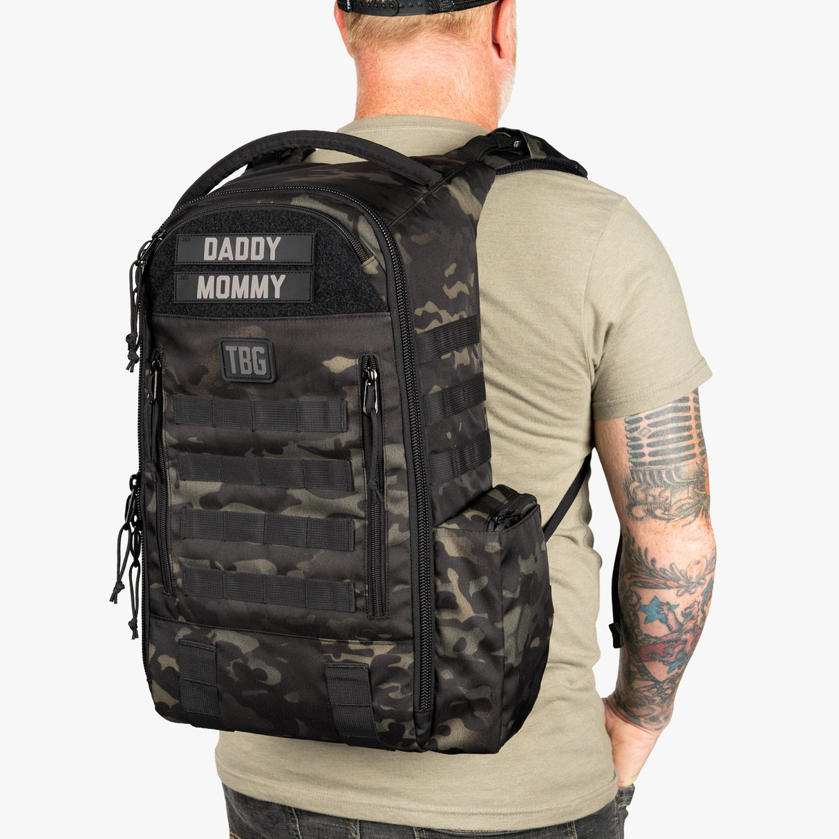 Black camouflage daypack diaper bag with baby carrier straps, labeled with "Daddy," "Mommy," and "TBG" patches, worn by a man.