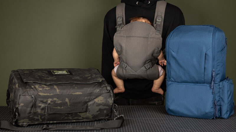 Adult in a black hoodie wearing a gray baby carrier with a baby, flanked by a camouflage duffel bag on the left and a large blue backpack on the right, against an olive-green background.