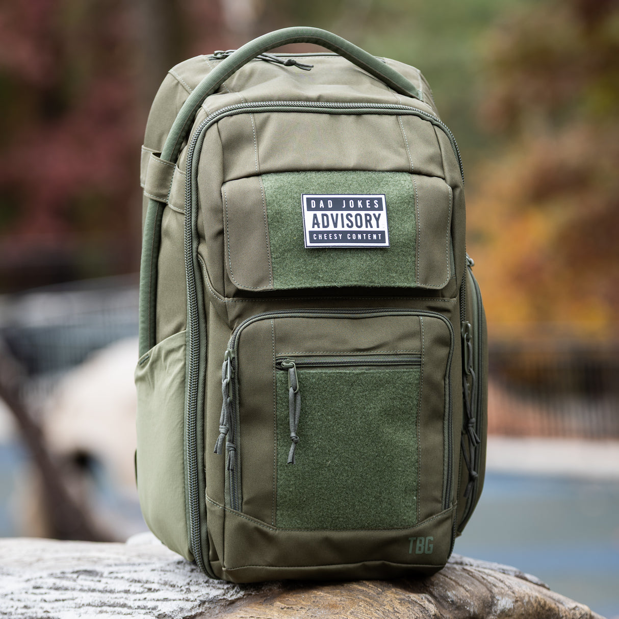 Olive green backpack featuring a "Dad Jokes Advisory" patch, designed for rugged parenting adventures.