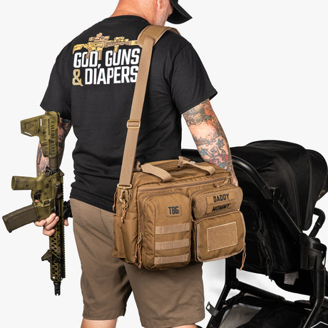 Man wearing a black t-shirt with 'Guns & Diapers' text, holding a rifle, and carrying a tactical bag with a stroller in the background.