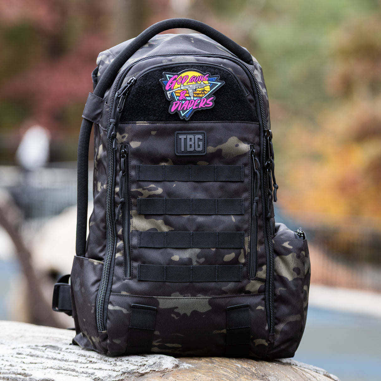 Tactical Baby Gear backpack featuring a colorful 80's style patch that reads "God, Guns & Diapers" on a camo background.
