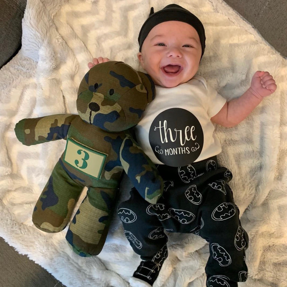 Baby wearing a 'three months' shirt with a camouflage teddy bear, lying on a textured blanket.