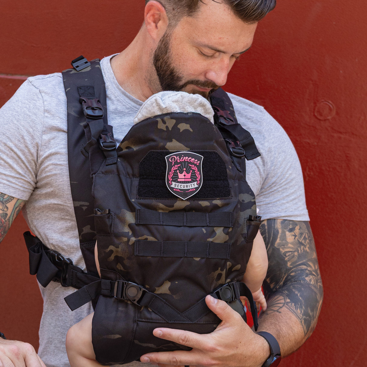 A dad wearing a camo baby carrier with a pink Princess Security patch, holding his child close.
