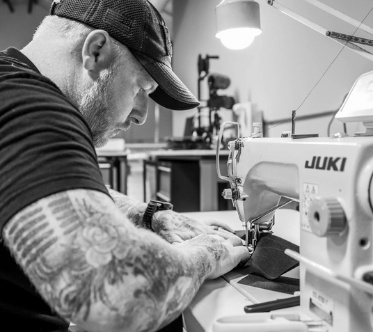 Black and white image of TBG founder Beav Brodie, focused on sewing fabric with a sewing machine in a workshop.