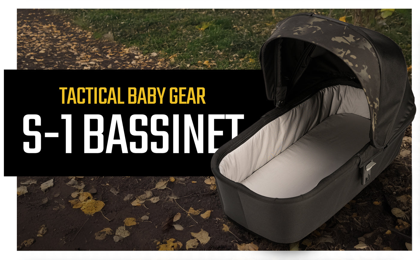 Camouflage patterned baby bassinet with gray trim, displayed outdoors on a leaf-strewn path.
