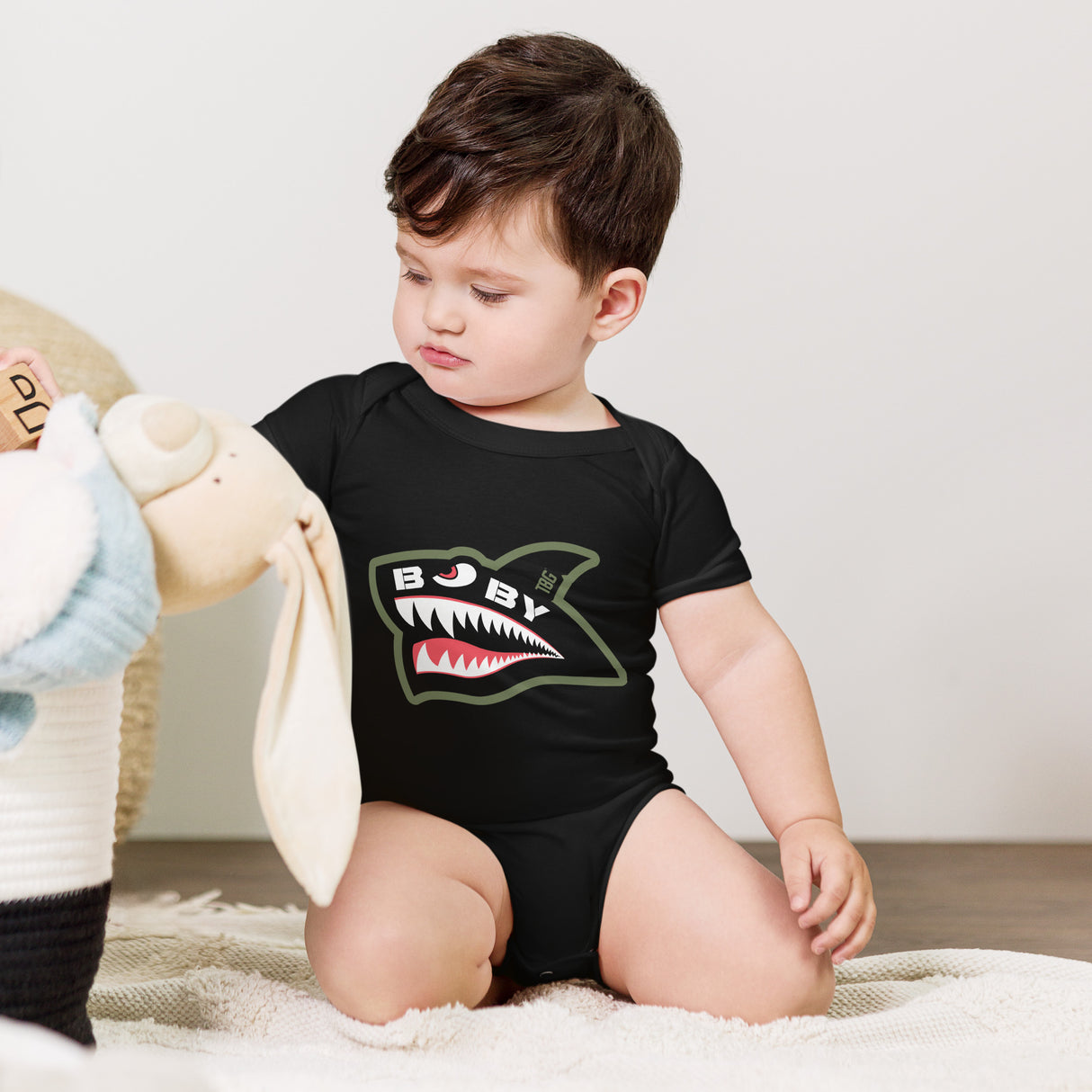 Baby wearing black onesie with a fierce, military-inspired shark logo in white, red, and green, perfect for rugged parenting.