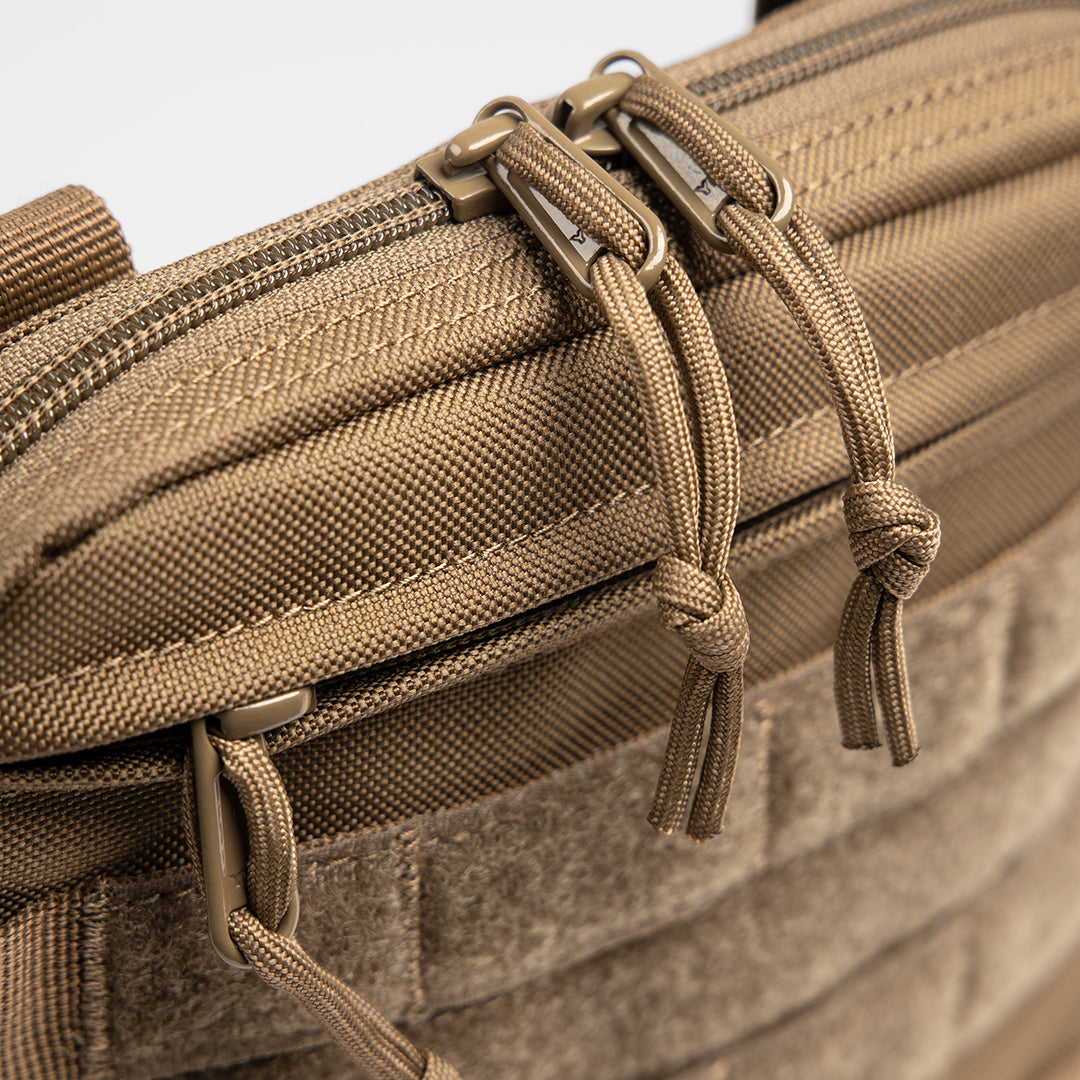 Close-up of a tan, rugged, military-inspired parenting bag with sturdy zippers, durable fabric, and knotted zipper pulls.