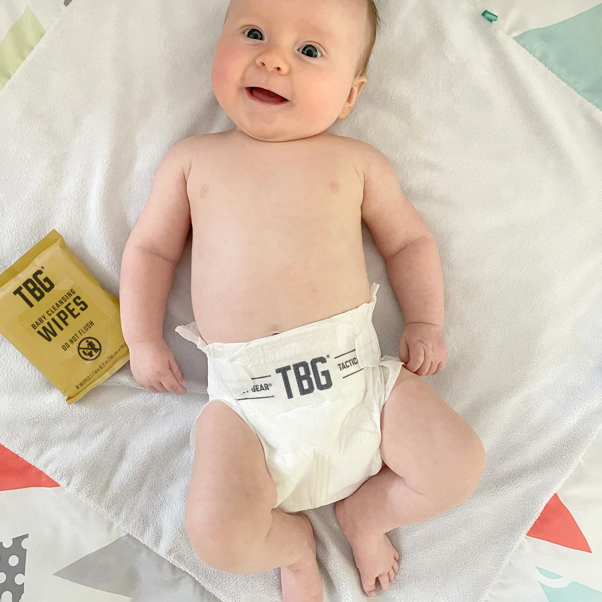 A happy baby lying on a colorful blanket, wearing a Tactical Baby Gear premium diaper, with wipes nearby.