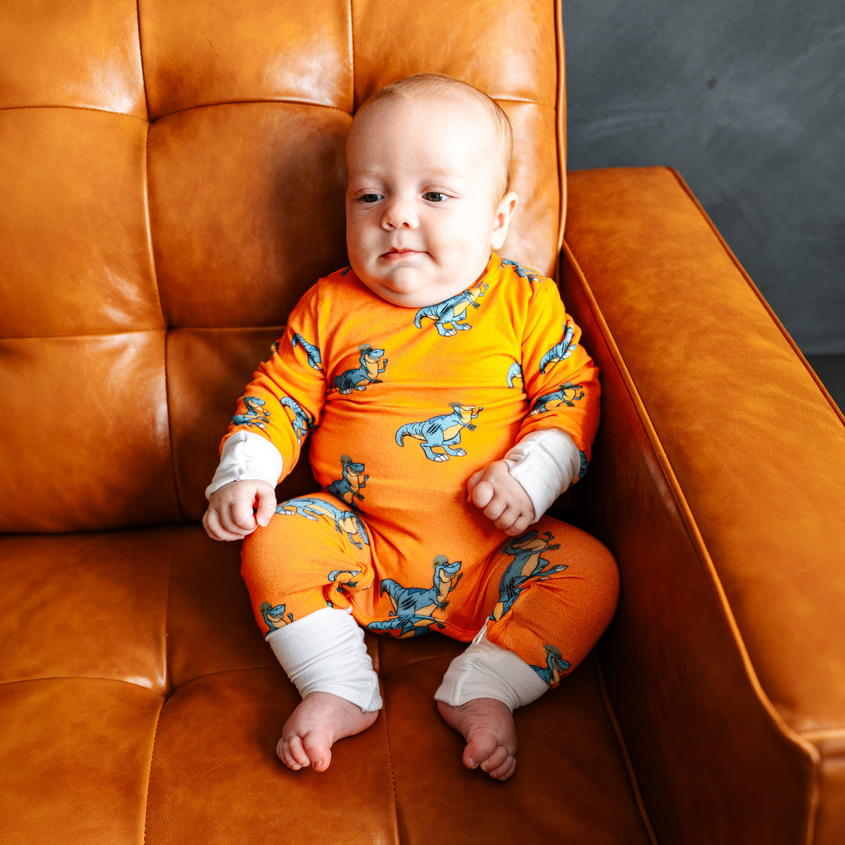 Infant in orange dinosaur-themed zippie pajamas sitting on a brown leather chair, looking content and relaxed.