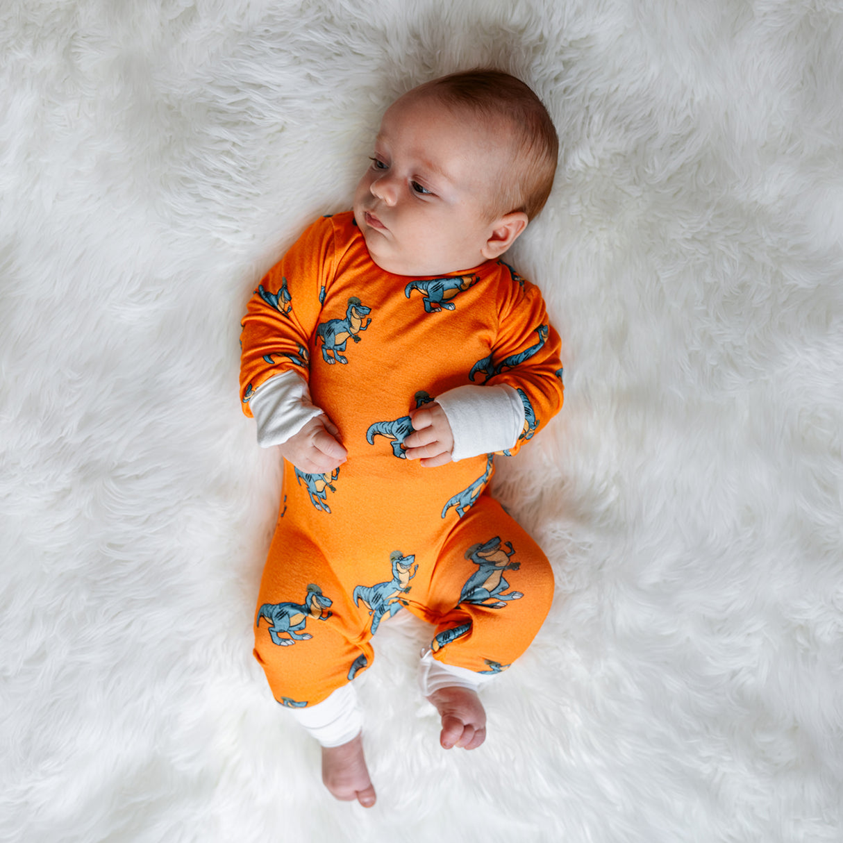 Infant in orange Dangerous Dino Zippie pajamas, lying on a fluffy white surface, looking thoughtfully to the side.