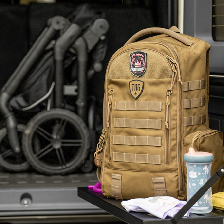Rugged tan military-inspired diaper backpack with patches, multiple compartments, and MOLLE straps for modern parents.