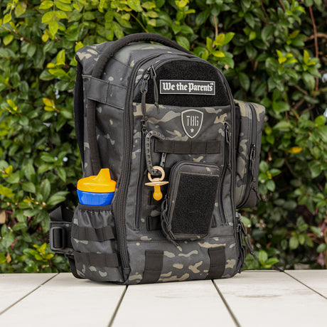 Military-inspired camouflage diaper bag with multiple zippered compartments, front patch, and side bottle holder.