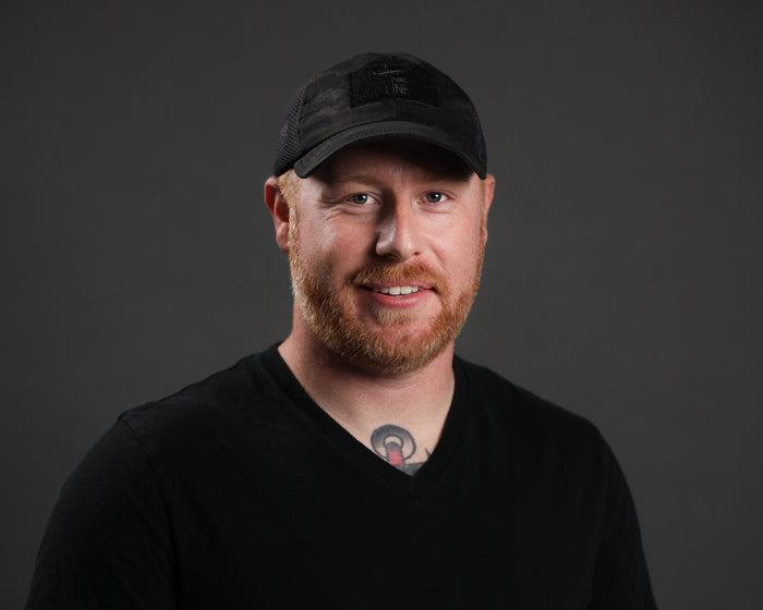 Man wearing a black tactical cap and black T-shirt, smiling against a grey background.