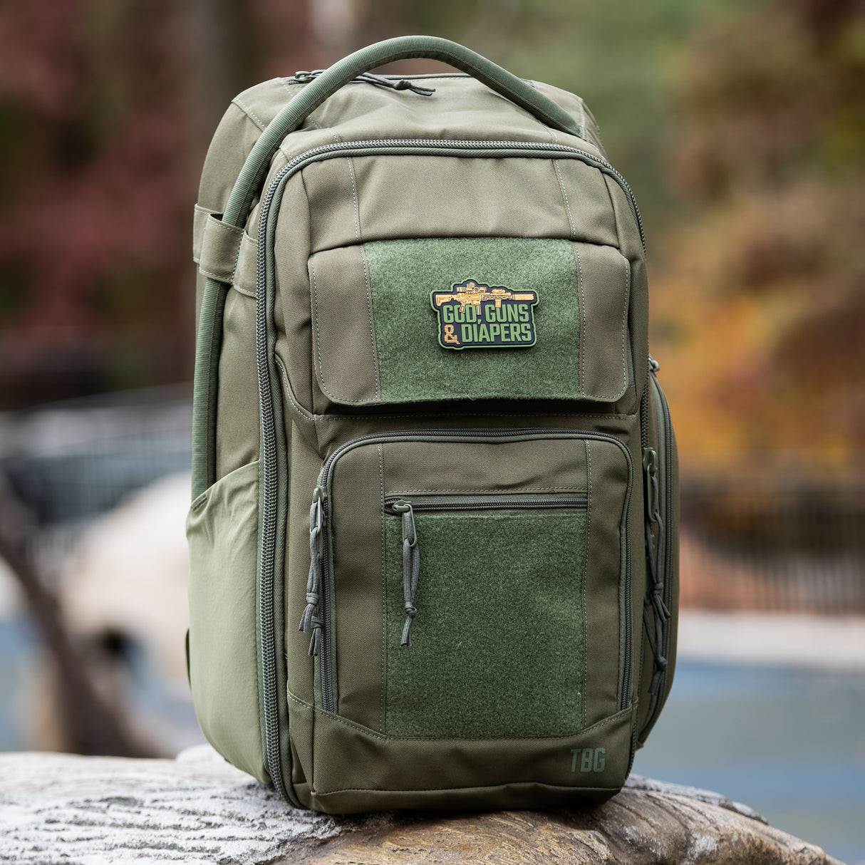 Olive green tactical backpack featuring a "God, Guns & Diapers" patch, designed for rugged parenting needs.