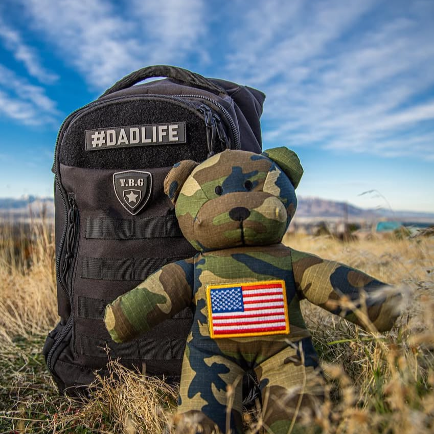 Camouflage teddy bear with tactical diaper bag and American flag patch in a field