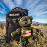 Camouflage teddy bear with tactical diaper bag and American flag patch in a field