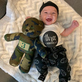 Baby wearing a 'three months' shirt with a camouflage teddy bear, lying on a textured blanket.