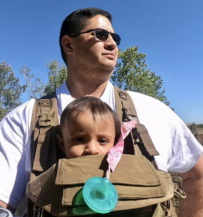 Man wearing a military-style baby carrier in olive green, with front pockets and a child peeking out.