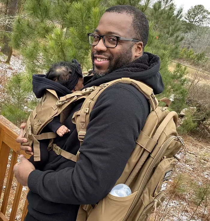 Man with glasses wearing a tan baby carrier and matching backpack, holding a baby, outdoors.