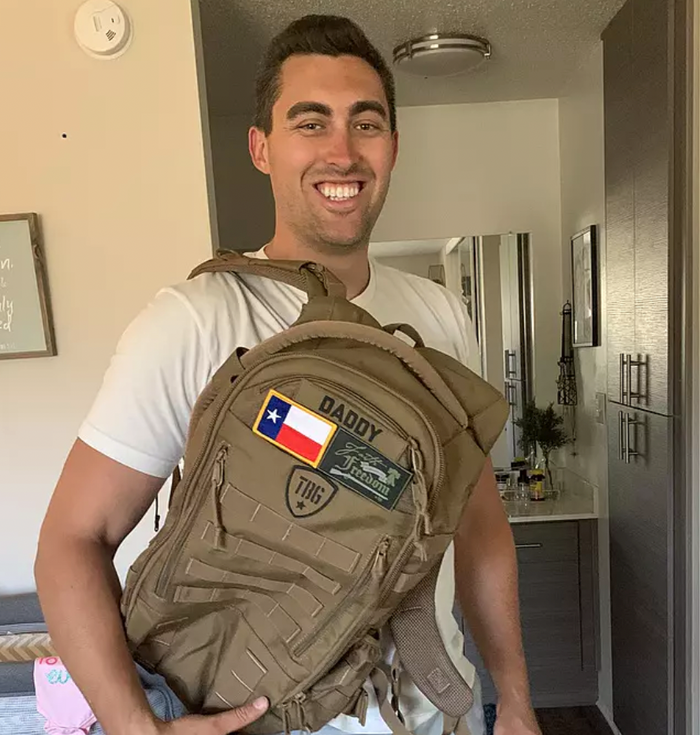 Man smiling, holding a child and wearing a tan military-style backpack with patches.
