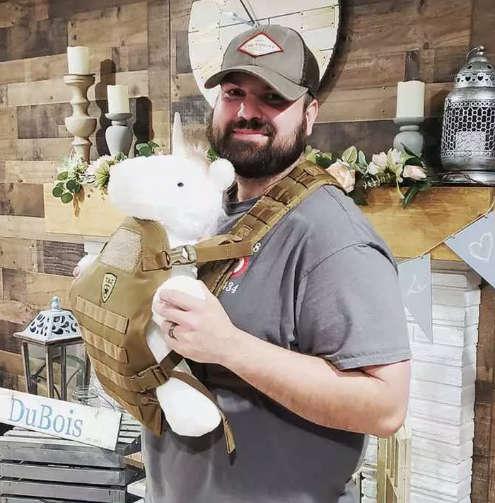 Man in cap holding a white plush bear, wearing a khaki tactical baby carrier.