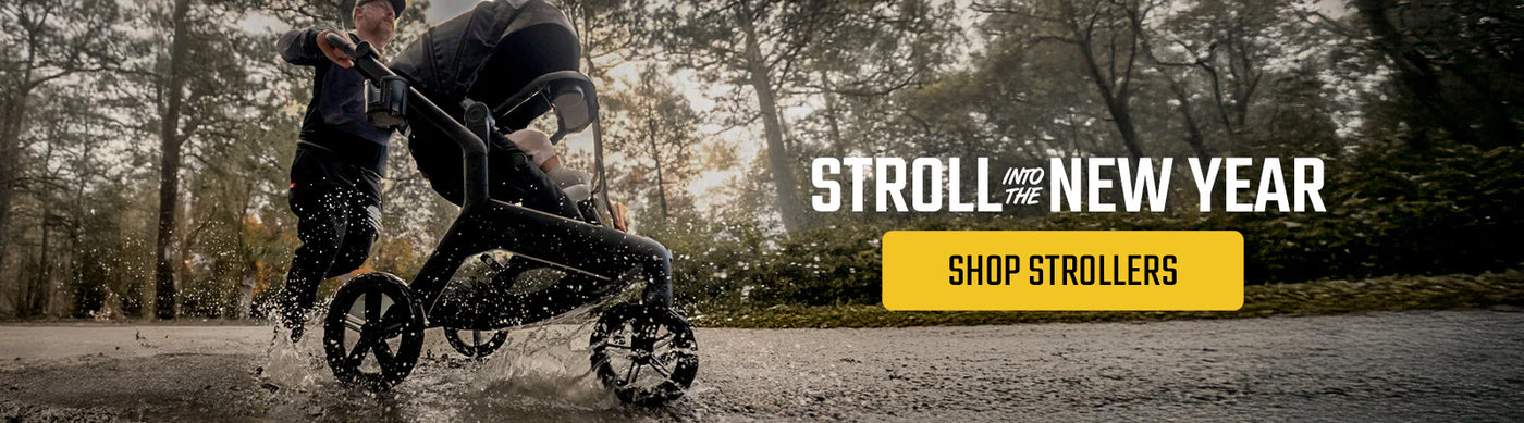 Man pushing a black stroller through a puddle on a forest path, with text reading “Stroll Into the New Year” and a yellow button labeled “Shop Strollers.”