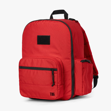 Alt text: Bright red classic diaper backpack with multiple compartments and zippers, featuring a black handle and side pocket detailing, and a black square logo patch on the front.