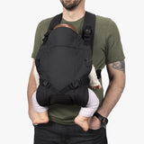 Alt text: Man wearing a black ergonomic baby carrier with adjustable straps, holding an infant securely. The carrier allows face-in position for enhanced bonding and comfort.