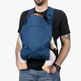 A man wearing a black T-shirt and jeans demonstrates a Baby Carrier Lite in blue, carrying a toddler with visible legs and arms hugged around his waist. The carrier features adjustable shoulder straps and secure buckles for support and comfort.