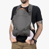 A man wearing a black shirt demonstrates a modern, gray baby carrier, securing a baby in front against his chest with adjustable padded straps and secure buckles.