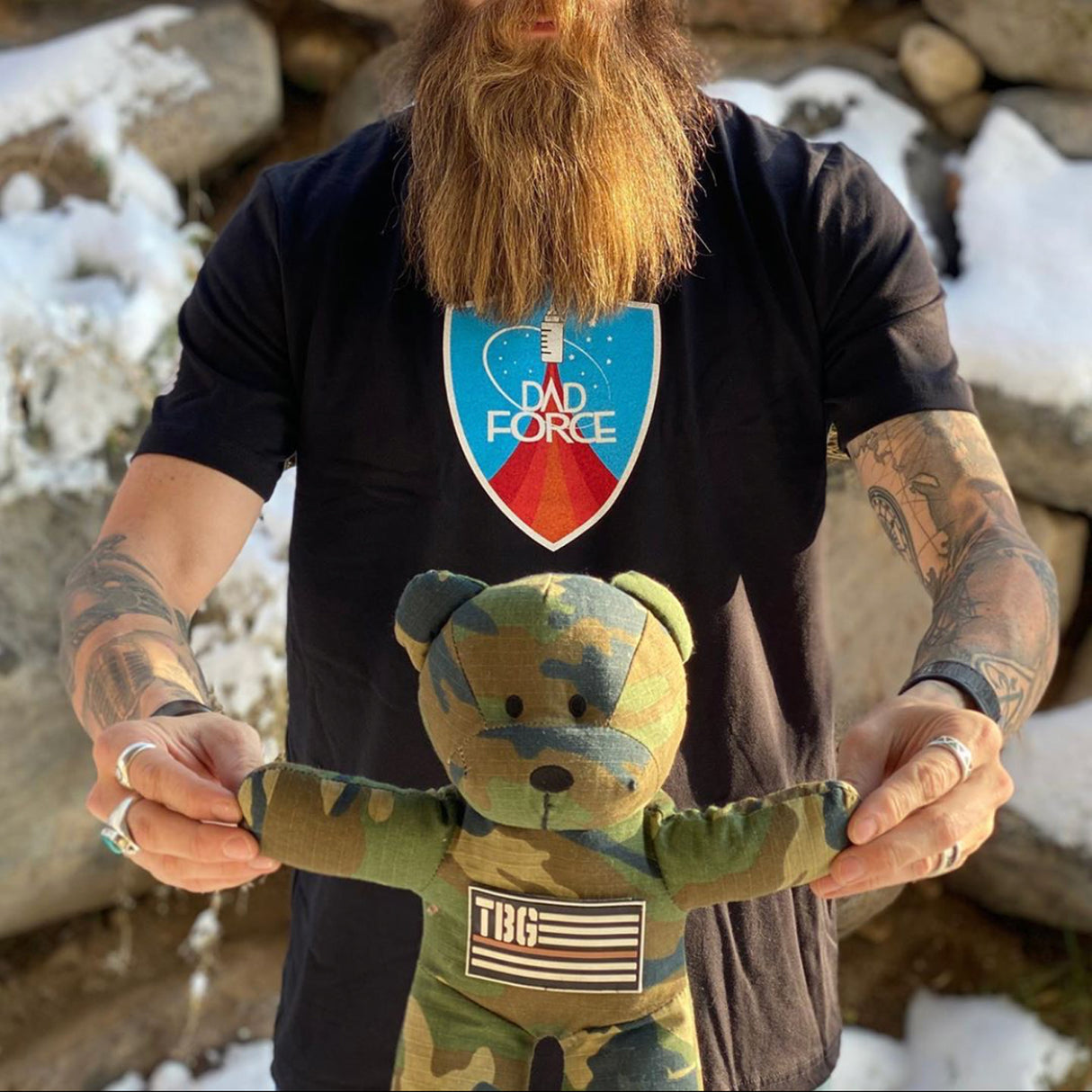 Dad holding a camouflage plush teddy bear in front of a rocky, snowy outdoor background. Rugged military-inspired parentin...